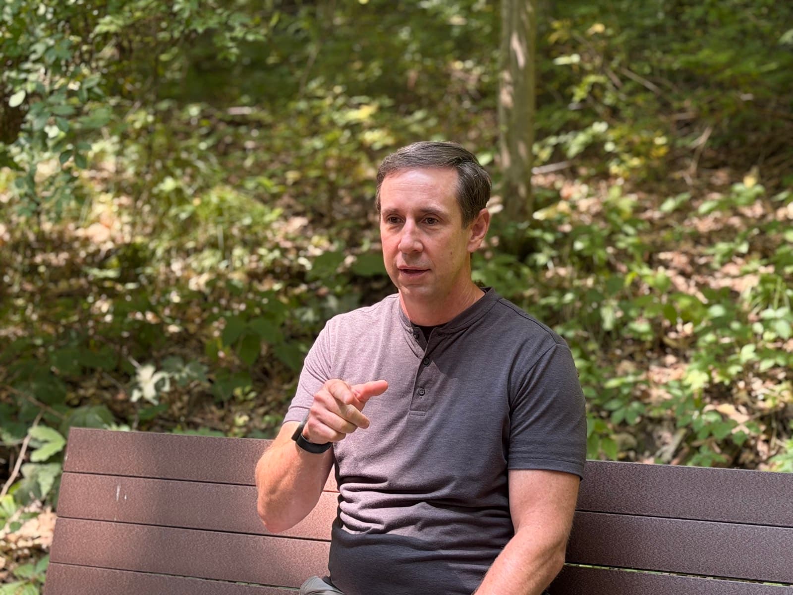 Gregg Huffman seated on a trailside bench, pointing at the camera mid-conversation
