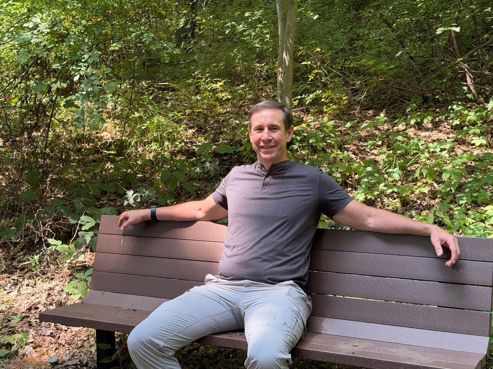 Gregg Huffman on a trailside bench, arms open in welcome, room on the bench beside him