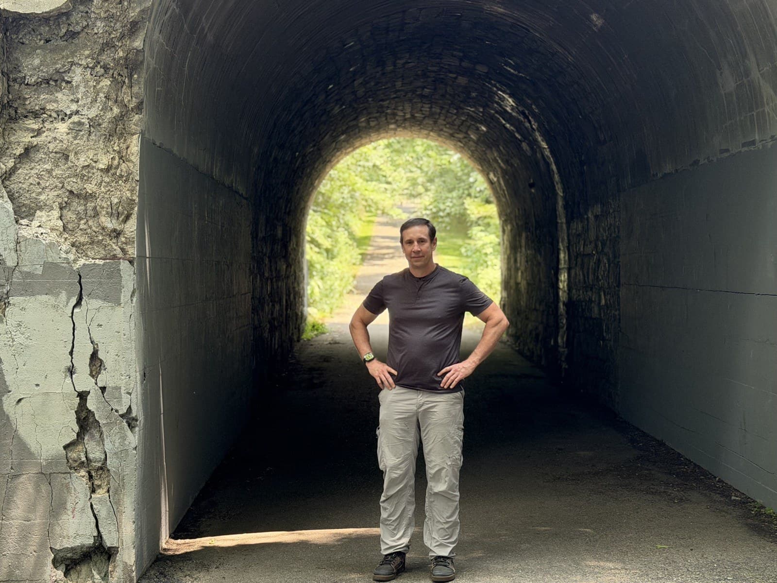 Gregg Huffman standing inside a dark stone tunnel, green light and the trail visible at the far end