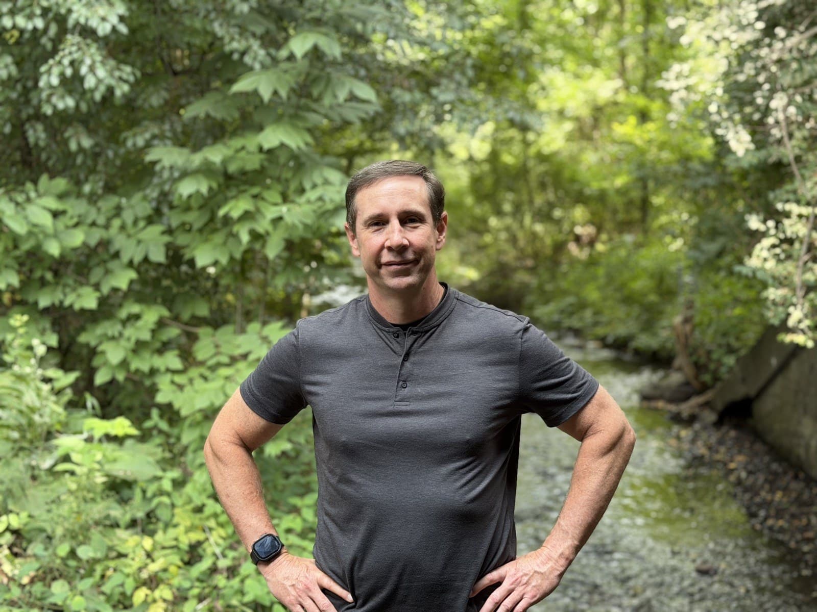 Gregg Huffman standing beside a creek in green Michigan woods, hands on his hips, facing the camera